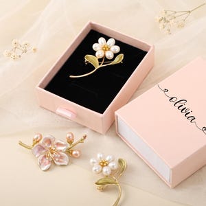 Puede incluir: Una caja de regalo rosa claro con el nombre olivia impreso en la tapa, que contiene un broche de flor dorado con p&eacute;talos de perlas blancas. Dos broches de flores adicionales, uno con p&eacute;talos de esmalte rosa y otro con p&eacute;talos de perlas blancas, yacen cerca.