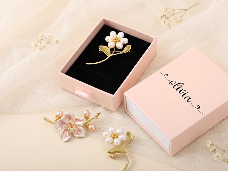 Puede incluir: Una caja de regalo rosa claro con el nombre olivia impreso en la tapa, que contiene un broche de flor dorado con p&eacute;talos de perlas blancas. Dos broches de flores adicionales, uno con p&eacute;talos de esmalte rosa y otro con p&eacute;talos de perlas blancas, yacen cerca.