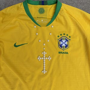 May include: A yellow Brazil soccer jersey with a green collar and cuffs. The jersey features the Nike logo, the CBF crest, and a pearl cross design. The jersey has a V-neck and is made of a breathable material.