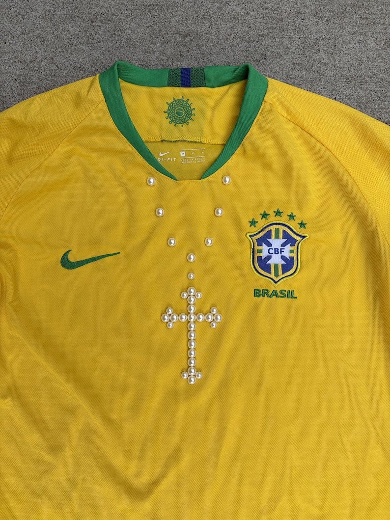 May include: A yellow Brazil soccer jersey with a green collar and cuffs. The jersey features the Nike logo, the CBF crest, and a pearl cross design. The jersey has a V-neck and is made of a breathable material.