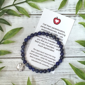 May include: A blue lapis lazuli beaded bracelet with a silver apple charm. A card with text about appreciation for educators is next to the bracelet. Green leaves are in the background.