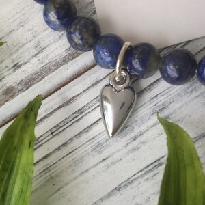 May include: A beaded bracelet featuring dark blue lapis lazuli stones. A silver heart charm hangs from the bracelet. The bracelet is displayed on a white wooden surface with green leaves in the foreground.