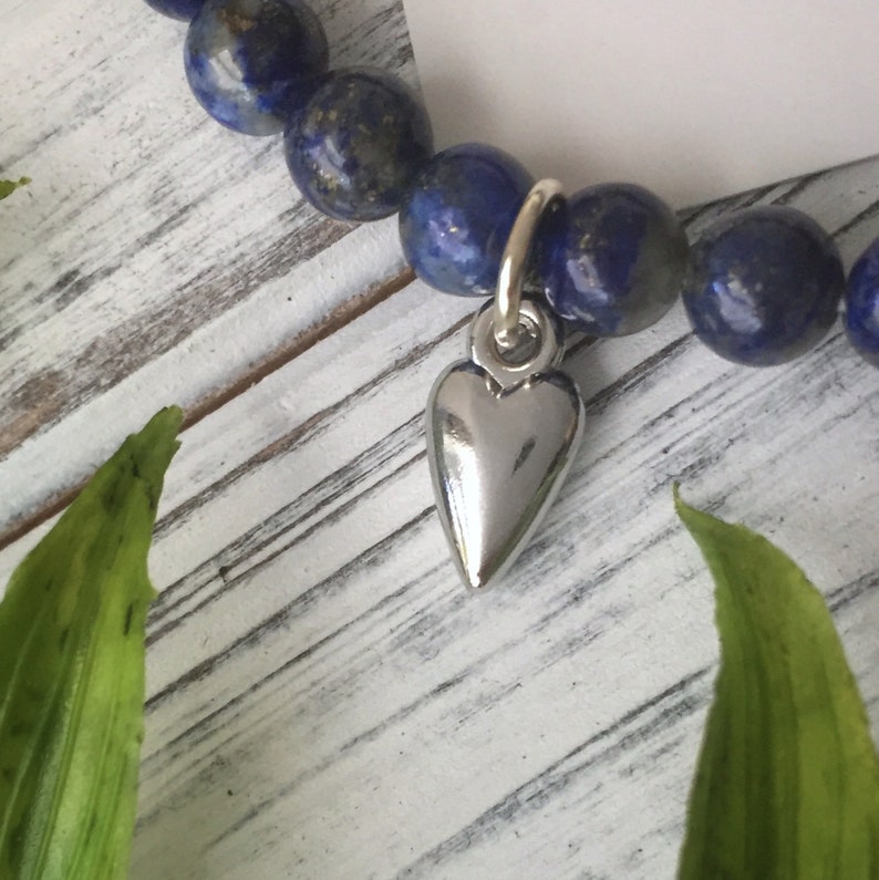 May include: A beaded bracelet featuring dark blue lapis lazuli stones. A silver heart charm hangs from the bracelet. The bracelet is displayed on a white wooden surface with green leaves in the foreground.