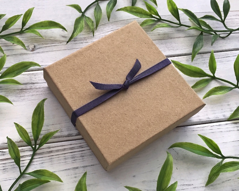 May include: A small, rectangular, brown cardboard gift box tied with a dark blue ribbon. The box is closed with a bow. The background is a white wooden surface with green leaves.
