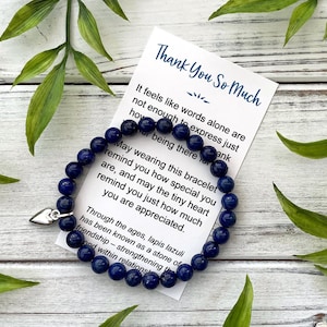 May include: A dark blue lapis lazuli bead bracelet with a silver heart charm. The bracelet is next to a card that says "Thank You So Much". Green leaves are in the background.