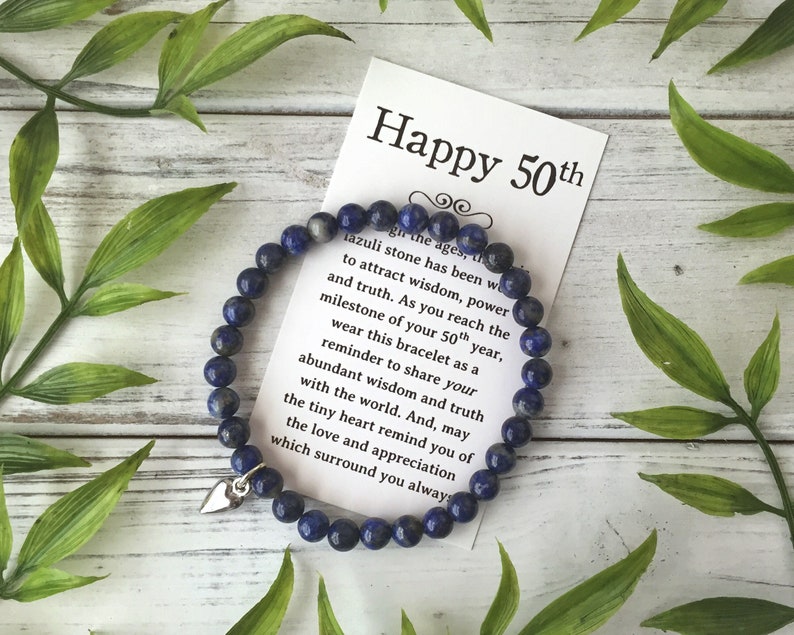 May include: A dark blue beaded bracelet with a silver heart charm, displayed with a card that reads "Happy 50th". The card also includes text about wisdom and truth. Green leaves surround the bracelet.