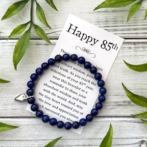 Peut inclure: Un bracelet de perles bleu foncé avec un charm en forme de cœur argenté repose sur une surface en bois blanc. Une carte avec le texte "Happy 85th" et un message sur la sagesse et la vérité est à côté du bracelet. Des feuilles vertes sont éparpillées.