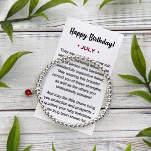 May include: A silver beaded bracelet with a red charm, presented with a card that reads "Happy Birthday!" and "July." The card includes a message about the bracelet's symbolism and the qualities of the ruby birthstone.