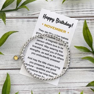 May include: A silver beaded bracelet with a small yellow charm, displayed with a card that reads "Happy Birthday! NOVEMBER." The card includes text about November babies. Green leaves are arranged around the bracelet and card.