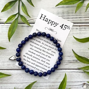 May include: A dark blue beaded bracelet with a silver heart charm, resting on a white card that reads "Happy 45th." The card is surrounded by green leaves, creating a natural aesthetic. The bracelet is made of small, round beads.