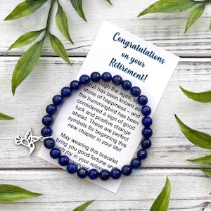May include: A circular bracelet made of deep blue lapis lazuli beads featuring a silver-toned hummingbird charm. The bracelet rests on a white card printed with the text Congratulations on your Retirement and a message about the stone and bird.