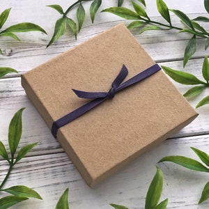 May include: A small, rectangular, brown cardboard gift box tied with a dark blue ribbon. The box is closed with a bow. The background is a white wooden surface with green leaves.