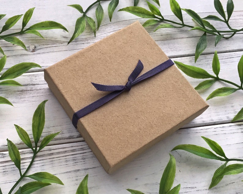 May include: A small, rectangular, brown cardboard gift box tied with a dark blue ribbon. The box is closed with a bow. The background is a white wooden surface with green leaves.