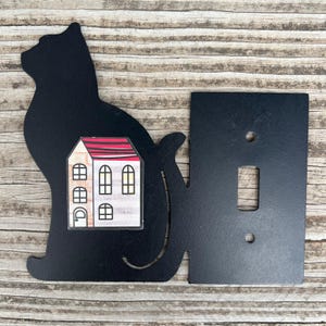 以下が含まれることがあります： 黒い金属製の猫型ライトスイッチカバー。赤い屋根と黒い窓枠のある白い壁の家のグラフィック。カバーには標準的なライトスイッチの開口部とネジ穴が含まれています。