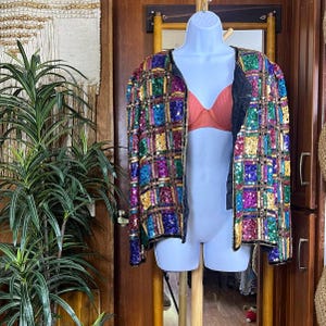 May include: A vibrant sequined jacket featuring a grid design of colourful squares in shades of blue, green, purple, and gold. The jacket has black trim and is displayed on a white mannequin. The jacket is open, revealing a peach-coloured bra.