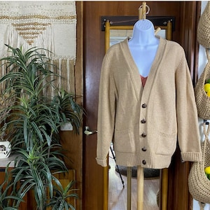 Op de afbeelding: Een beige vest met een V-hals, knoopsluiting en twee voorzakken. De trui wordt op een mannequin gepresenteerd. De mouwen zijn opgerold. De achtergrond omvat een houten deur, een spiegel en een plant.