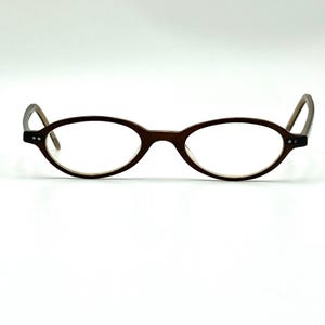 May include: A pair of oval-shaped eyeglasses with a dark brown frame. The glasses feature thin rims and two small silver-toned rivets on each side of the front frame. The temples are curved and extend outward from the sides of the frame.