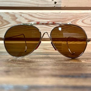 Könnte beinhalten: Sonnenbrille im Aviator-Stil mit goldfarbenem Metallrahmen und braunen Gläsern. Der obere Steg hat ein rot-weißes Muster. Klassisches Design, passend für verschiedene Stile. Durchmesser der Gläser: ca. 5 cm.