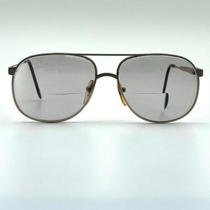 May include: Aviator-style spectacles featuring a slim, dark metallic frame and a double bridge. The lenses are clear and include a distinct horizontal line across the lower section, denoting bifocal lenses. The nose pads are a translucent amber colour.