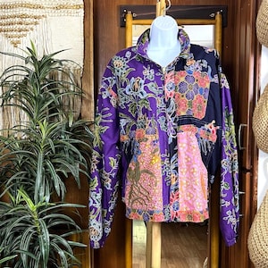 May include: A vintage zip-up jacket with a vibrant floral pattern in purple, pink, and blue. The jacket features a high collar, front pockets, and a mix of patterned panels. The jacket is displayed on a white mannequin.