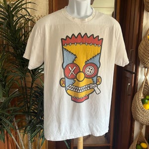 May include: Cream-colored t-shirt featuring a cartoon face design. The design includes yellow, red, blue, and white elements, with a zipper and button details. The graphic is a stylized representation of a character.