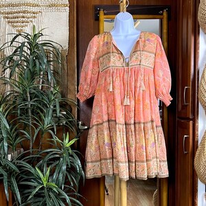 May include: A peach-coloured mini dress featuring a floral pattern in shades of pink, orange, and yellow. The dress has three-quarter length sleeves with elastic cuffs, a V-neckline with two hanging tassels, and a tiered skirt design.