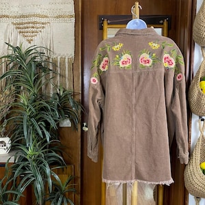 May include: A brown corduroy shirt-dress with long sleeves and a frayed hem. The back of the shirt features colorful floral embroidery in pink, yellow, and green. The shirt is hanging on a wooden rack.