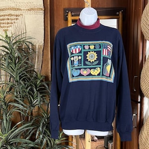 Puede incluir: Sudadera azul marino con cuello alto burdeos. La parte delantera presenta un colorido diseño de patchwork con corazones, una bandera estadounidense, un girasol y otros símbolos patrióticos. La parte superior de manga larga se muestra en un maniquí.