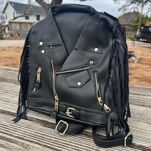 May include: Black faux leather jacket with gold zippers, buttons, and buckle details. The jacket features fringe accents on the sleeves and a belt at the waist. The jacket has a classic biker style.