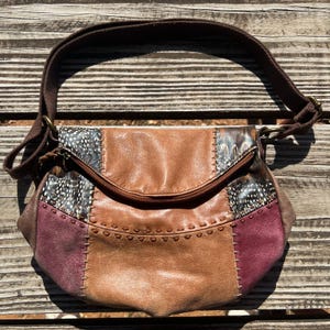 Könnte beinhalten: Eine braune Leder-Umhängetasche mit Patchwork-Design. Die Tasche hat eine Mischung aus braunen, beigen und bordeauxfarbenen Lederpaneelen, mit Federakzenten und einem braunen Stoffriemen. Die Tasche hat einen Reißverschluss und Metallbeschläge.