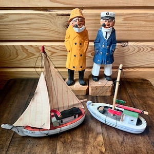 Pode incluir: Estatuetas náuticas e veleiros de madeira. As estatuetas retratam um pescador com um casaco amarelo e um capitão com um casaco azul. Também estão presentes dois pequenos veleiros, um com uma faixa vermelha e outro com detalhes verdes e vermelhos. Os veleiros têm o texto "Cape Cod".