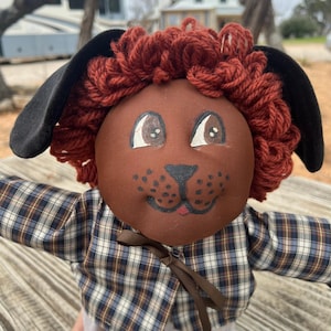May include: A handcrafted doll with a brown face, red yarn hair, and black floppy ears. The doll has painted eyes and a smiling mouth. It is wearing a plaid shirt with a brown ribbon tie.
