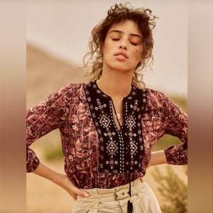 May include: A long-sleeved blouse with a burgundy and cream floral pattern. The blouse features a black embroidered yoke with geometric designs and a tassel detail at the neckline. The model is wearing khaki-colored pants.