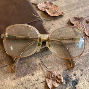 May include: Vintage eyeglasses with a light brown frame and clear lenses. The glasses have a rounded shape and gold-colored metal accents. The image is set on a wooden surface with dried leaves and a brown leather case.