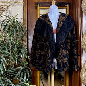 May include: A long-sleeved jacket featuring a black and bronze-colored floral pattern throughout. The garment includes a V-neckline and long black fringe detailing that runs along the front lapels and down the center of the jacket body.
