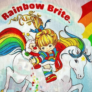 May include: A vintage Rainbow Brite graphic featuring the character riding a white horse with a rainbow mane. The image includes the text "Rainbow Brite" in red, with a rainbow and stars.