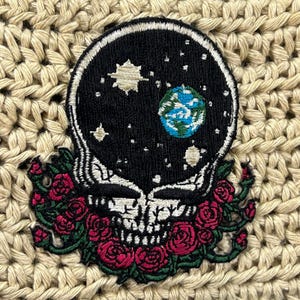 May include: An embroidered patch featuring a black skull silhouette filled with a starry night sky, a glowing sun, and a blue and green Earth. The base of the skull is surrounded by a cluster of red roses with green leaves and stems.