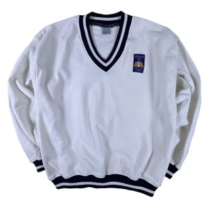 Puede incluir: Sudadera blanca con cuello en V y ribete a rayas azul marino y blanco en el cuello, los puños y el dobladillo. Cuenta con un parche con el logotipo de los Juegos Olímpicos de Atlanta 1996. Manga larga, corte holgado.