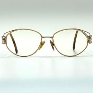 May include: Gold-tone metal eyeglasses with oval-shaped frames. The temples feature a twisted wire texture, and the hinges are accented with silver-tone scrollwork. The nose pads are gold-tone, and the temple tips are a brown tortoiseshell pattern.
