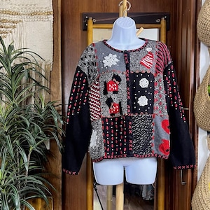 Könnte beinhalten: Ein Patchwork-Cardigan in Schwarz, Grau und Rot mit weihnachtlichen Applikationen. Der Pullover hat einen runden Ausschnitt und eine Knopfleiste vorne. Die Ärmel und Kanten sind mit roten Nähten versehen.