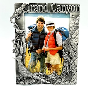 May include: A rectangular pewter picture frame featuring the embossed text Grand Canyon at the top. The frame is decorated with a lizard, a bird, cacti, a roadrunner, and a trail scene with figures on mules, surrounding a photo of two hikers.