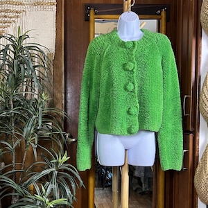 May include: A bright lime green long-sleeve cardigan with a textured, fuzzy fleece-like finish. The garment features a round neckline and a front button closure with four large, matching fabric-covered spherical buttons down the center.