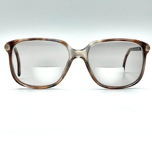 May include: Bifocal eyeglasses featuring a brown tortoiseshell frame with a translucent bridge and gold-tone metal accents at the hinges. The lenses are clear with a visible horizontal line separating the upper and lower segments of the bifocal design.