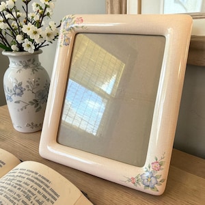 May include: A rectangular, light pink picture frame with rounded corners. The frame is decorated with small floral clusters in pink, blue, and green. A vase with white flowers and a book are in the background.