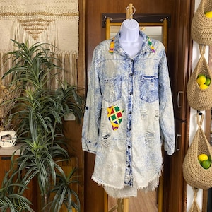 May include: Acid wash denim shirt dress with distressed details. Features colorful geometric patches and a frayed hem. The long-sleeved shirt has a button-down front and a collar with colorful trim.