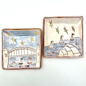 May include: Two square ceramic dishes with hand-painted designs. One dish features the Sydney Harbour Bridge over water with sailboats and yellow stars. The other dish depicts the Sydney Opera House, sailboats, fish, and yellow stars on a light background.