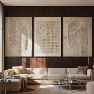 May include: Three large, textured wall art pieces with cream-colored, abstract designs hang above a cream-colored sectional sofa. The art features curved and grid-like patterns. A round, glass-topped coffee table and decorative pillows complete the living room setting.