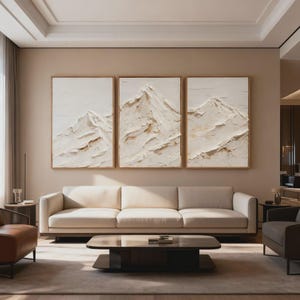 Puede incluir: Tres pinturas abstractas enmarcadas de montañas con pintura blanca y beige texturizada. La obra de arte cuelga sobre un sofá color crema en un salón moderno. Una mesa de centro de madera oscura y una alfombra de tonos neutros completan el espacio.