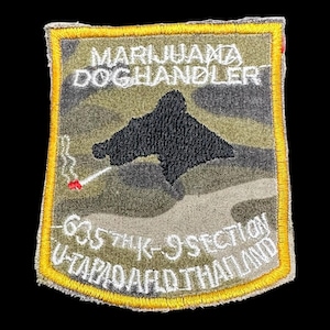 Könnte beinhalten: Gestickter Aufnäher mit Tarnmuster, mit dem Text "MARIJUANA DOGHANDLER" und "635TH K-9 SECTION U-TAPAO AIRFLD THAILAND". Eine schwarze Silhouette eines Hundes befindet sich in der Mitte, mit einer brennenden Zigarette. Der Aufnäher hat einen gelben Rand.