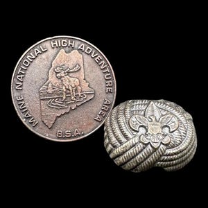 Op de afbeelding: Twee vintage Boy Scout-spelden. Eén is een ronde, zilverkleurige munt met de tekst "MAINE NATIONAL HIGH ADVENTURE AREA B.S.A." en een afbeelding van een eland. De andere is een zilverkleurige knoop met een lelie- en adelembleem.
